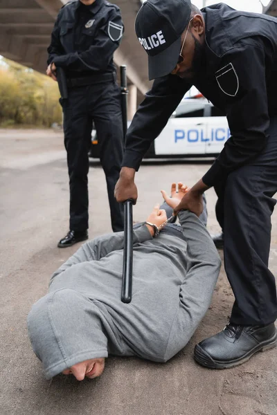 Agente di polizia afroamericano con manganello che arresta un criminale incappucciato sdraiato per strada su sfondo sfocato — Foto stock