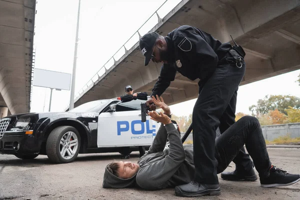 Full length of african american policeman aiming with pistol at scared offender lying on urban street on blurred background — Stock Photo