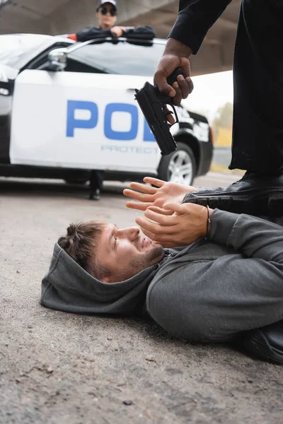 Policía afroamericana apuntando con pistola a delincuente asustado tendido en la calle con patrulla borrosa en el fondo - foto de stock