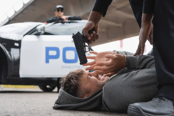 Policía afroamericano apuntando con pistola a delincuente asustado tendido en la calle sobre fondo borroso - foto de stock