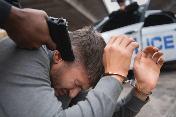 Vista de cerca del policía afroamericano apuntando con pistola al delincuente asustado sobre fondo borroso al aire libre - foto de stock