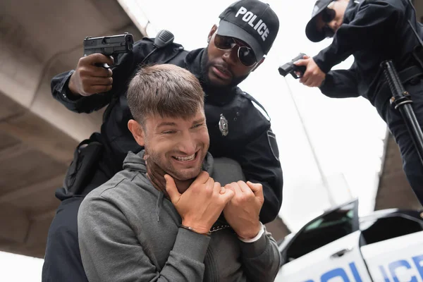 Low angle view of african american policeman choking offender while aiming with pistol near colleague on blurred background outdoors — Stock Photo