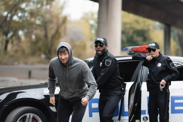 Hooded offender running from shocked multicultural police officers on blurred background on urban street — Stock Photo