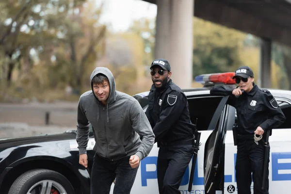 Ofensor encapuzado que foge de policiais multiculturais chocados em fundo embaçado na rua urbana — Fotografia de Stock