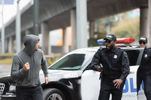 Delincuente encapuchado huyendo de agentes de policía multiculturales cerca de patrulla sobre fondo borroso en la calle urbana - foto de stock