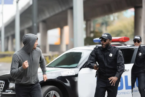 Criminoso encapuzado que foge de policiais multiculturais perto de carro de patrulha em fundo embaçado na rua urbana — Fotografia de Stock