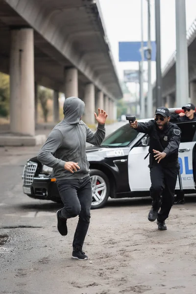 Full length of hooded offender running from shocked african american policeman aiming with pistol on blurred background on urban street — Stock Photo