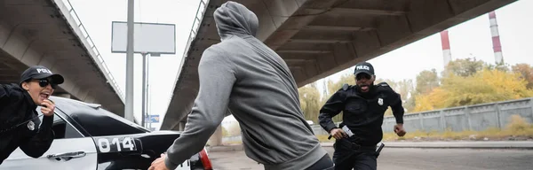 Delincuente encapuchado que huye de agentes de policía multiculturales cerca de coche patrulla en la calle urbana, pancarta - foto de stock