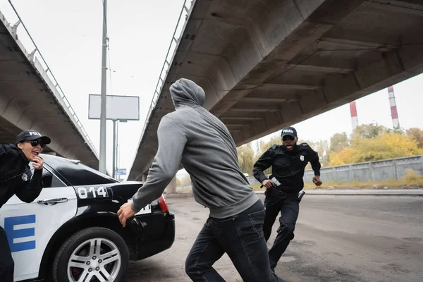 Ofensor encapuzado que foge de policiais multiculturais perto de carro de patrulha na rua urbana — Fotografia de Stock