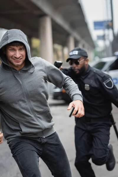 Hooded offender running from african american police officer aiming with gun on blurred background outdoors — Stock Photo