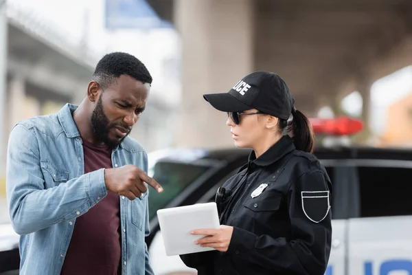 Worried african american victim pointing with finger at tablet in hands of policewoman on blurred background outdoors — Stock Photo