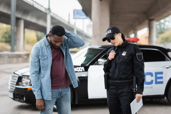 Policewoman talking on radio set near african american victim with blurred patrol car on background outdoors — Stock Photo
