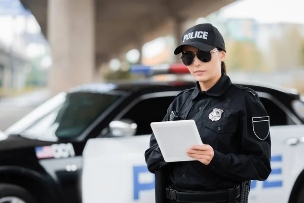 Young policewoman with tablet looking at camera with blurred patrol car on background outdoors — Stock Photo