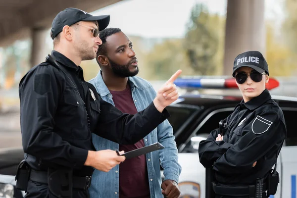 Africano americano vítima olhando para longe perto policial apontando com o dedo no fundo borrado ao ar livre — Fotografia de Stock