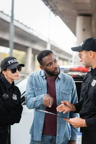 Victime afro-américaine pointant du doigt le presse-papiers tout en parlant avec un policier sur fond flou à l'extérieur — Photo de stock