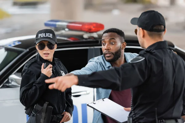 Conmocionada víctima afroamericana mirando hacia otro lado cerca de los oficiales de policía en el fondo borroso al aire libre - foto de stock