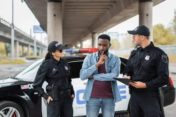 Policiais olhando para a vítima afro-americana pensando perto de carro de patrulha em fundo embaçado ao ar livre — Fotografia de Stock