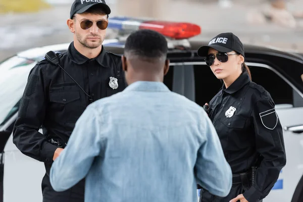 Vista posterior de la víctima afroamericana de pie cerca de oficiales de policía serios en un primer plano borroso al aire libre - foto de stock