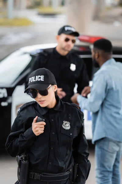 High angle view of policewoman talking on radio set with blurred colleague and african american man on background outdoors — Stock Photo