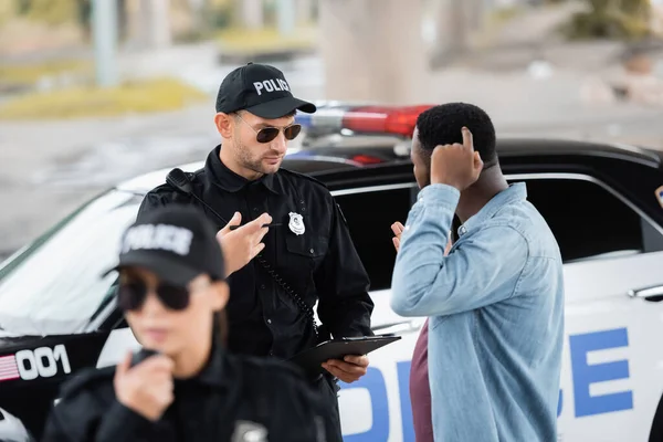 Policial conversando com afro-americano vítima perto de carro de patrulha com colega turvo em primeiro plano ao ar livre — Fotografia de Stock