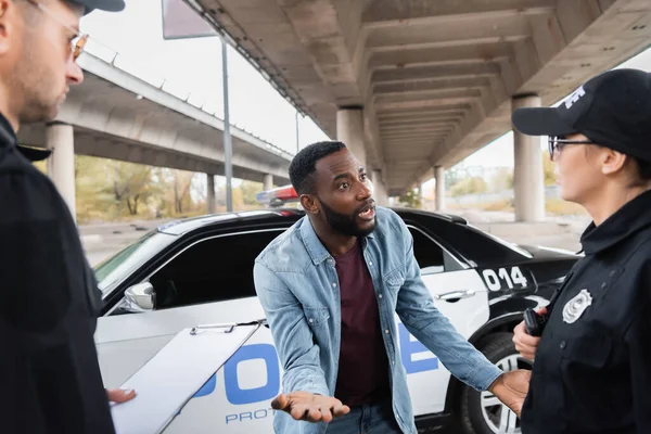 Irritated african american victim gesturing while arguing with police officers on blurred background on urban street — Stock Photo