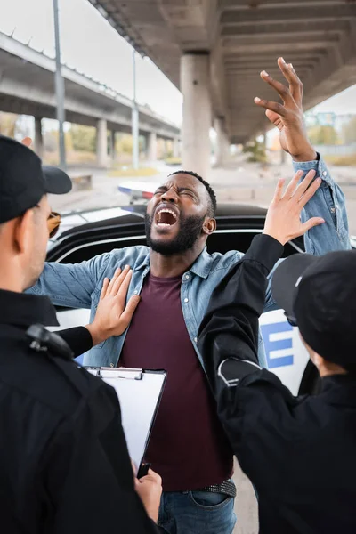 Back view of police officers calming despaired african american man near patrol car on blurred background on urban street — Stock Photo