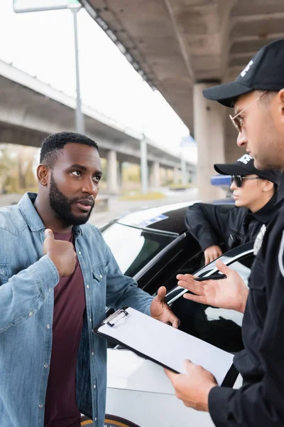 Víctima afroamericana hablando con policía sosteniendo portapapeles sobre fondo borroso al aire libre - foto de stock