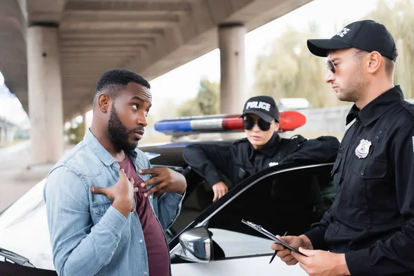 Victime afro-américaine irritée gestuelle tout en se disputant avec un policier avec une policière floue en arrière-plan à l'extérieur — Photo de stock