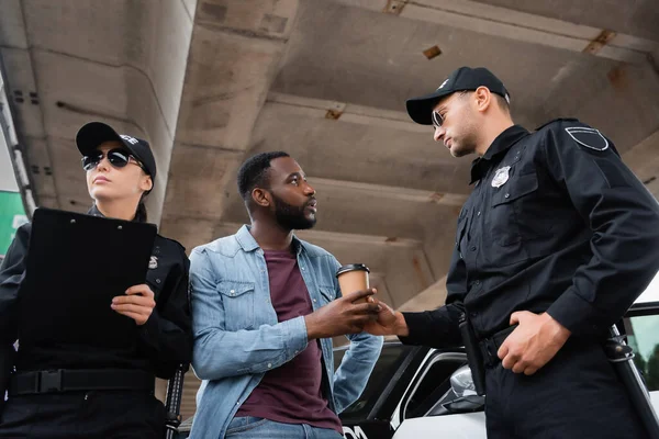 Low angle view of policeman giving coffee to go to african american victim near colleague with clipboard and car outdoors — Stock Photo