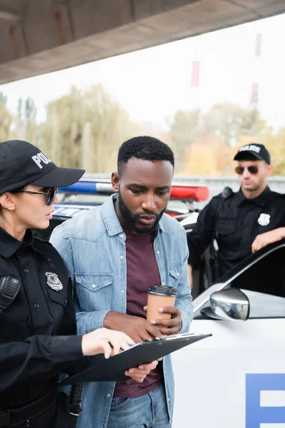Policía sosteniendo portapapeles cerca de víctima afroamericana con café para llevar y colega cerca de coche sobre fondo borroso - foto de stock
