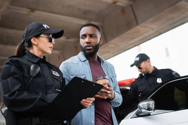 Policewoman with clipboard talking with african american victim with takeaway coffee near colleague and car on blurred background — Stock Photo