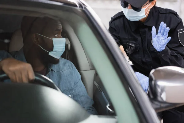 Poliziotta in lattice guanti saluto afroamericano conducente in maschera medica in auto in primo piano sfocato — Foto stock