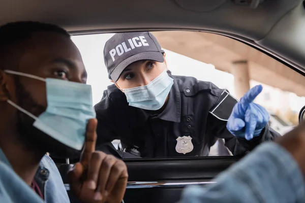 Mujer policía en máscara médica y guante de látex apuntando con el dedo cerca del conductor afroamericano sentado en el coche en primer plano borroso - foto de stock