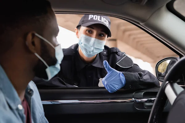 Police officer in medical mask and latex glove showing like to african american driver in auto on blurred foreground — Stock Photo