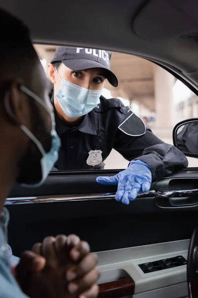 Поліцайка в медичній масці і латекс рукавичці дивлячись на африканського американця в автомобілі на розмитому передньому плані — стокове фото
