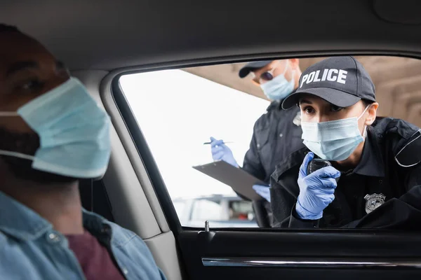 Policewoman in medical mask using walkie talkie near colleague with clipboard and african american driver on blurred foreground in car — Stock Photo