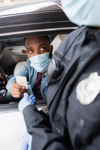 Conductor afroamericano en máscara médica dando licencia a policía en primer plano borroso cerca de coche - foto de stock