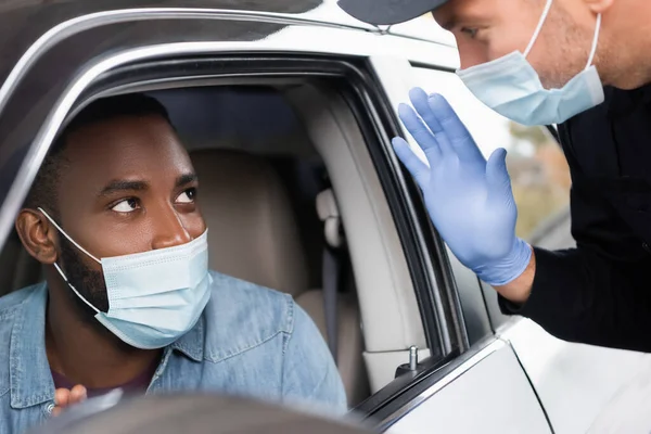 Oficial de policía en máscara médica y guante de látex saludando al conductor afroamericano en coche en primer plano borroso - foto de stock