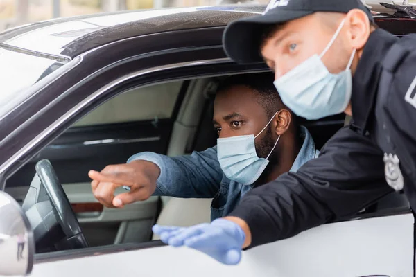 Conductor afroamericano en máscara médica señalando con el dedo cerca de oficial de policía en primer plano borroso cerca de coche - foto de stock