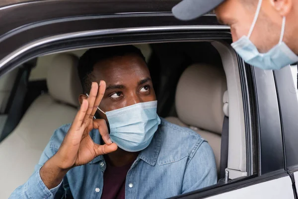 Conductor afroamericano en máscara médica mostrando un gesto aceptable a un policía cerca de auto en primer plano borroso - foto de stock