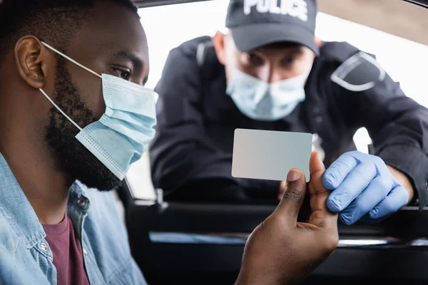 Conducteur afro-américain en masque médical titulaire d'un permis près de policier en gant de latex sur fond flou — Photo de stock