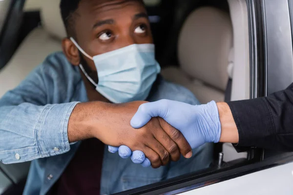 Police officer in latex glove handshaking with african american driver in medical mask on blurred background in car — Stock Photo