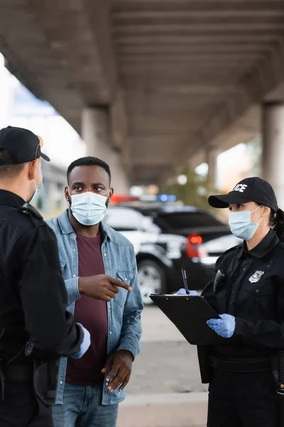 Víctima afroamericana en máscara médica señalando con el dedo cerca de agentes de policía con portapapeles en la calle urbana - foto de stock
