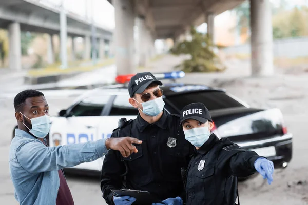 Africano americano vítima em máscara médica apontando com o dedo perto de policiais em luvas de látex com prancheta — Fotografia de Stock