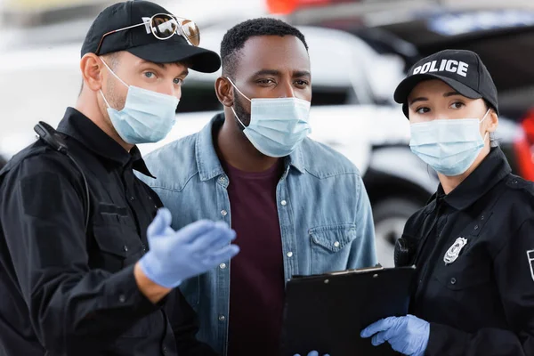 Víctima afroamericana en máscara médica parada cerca de agentes de policía con portapapeles en la calle urbana - foto de stock