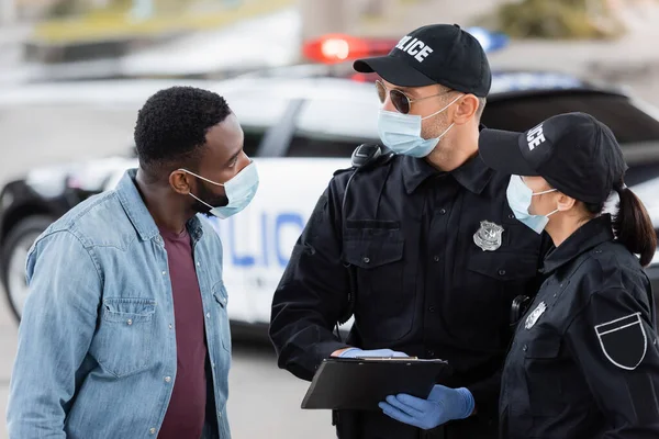Policías con máscaras médicas con portapapeles mirando a la víctima afroamericana en la calle urbana - foto de stock