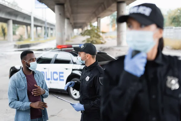 Африканская американская жертва в медицинской маске стоит рядом с офицером полиции и женщиной-полицейским на размытом переднем плане на городской улице — стоковое фото