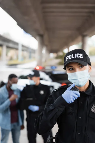 Policia en máscara médica usando walkie talkie cerca de víctima afroamericana y colega en fondo borroso en la calle urbana - foto de stock