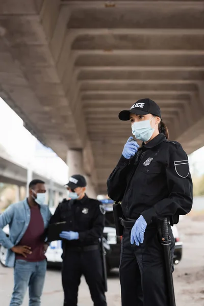 Policia con guantes de látex y máscara médica usando walkie talkie cerca de colega y víctima afroamericana sobre fondo borroso al aire libre - foto de stock