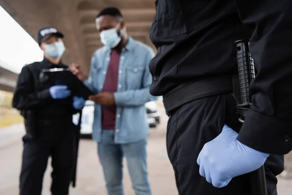 Ufficiale di polizia in guanto di lattice in piedi vicino afro-americano vittima e collega su sfondo sfocato all'aperto — Foto stock